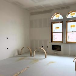 Angled view of a hotel room under construction. Arched double windows. Drywall and putty in place.